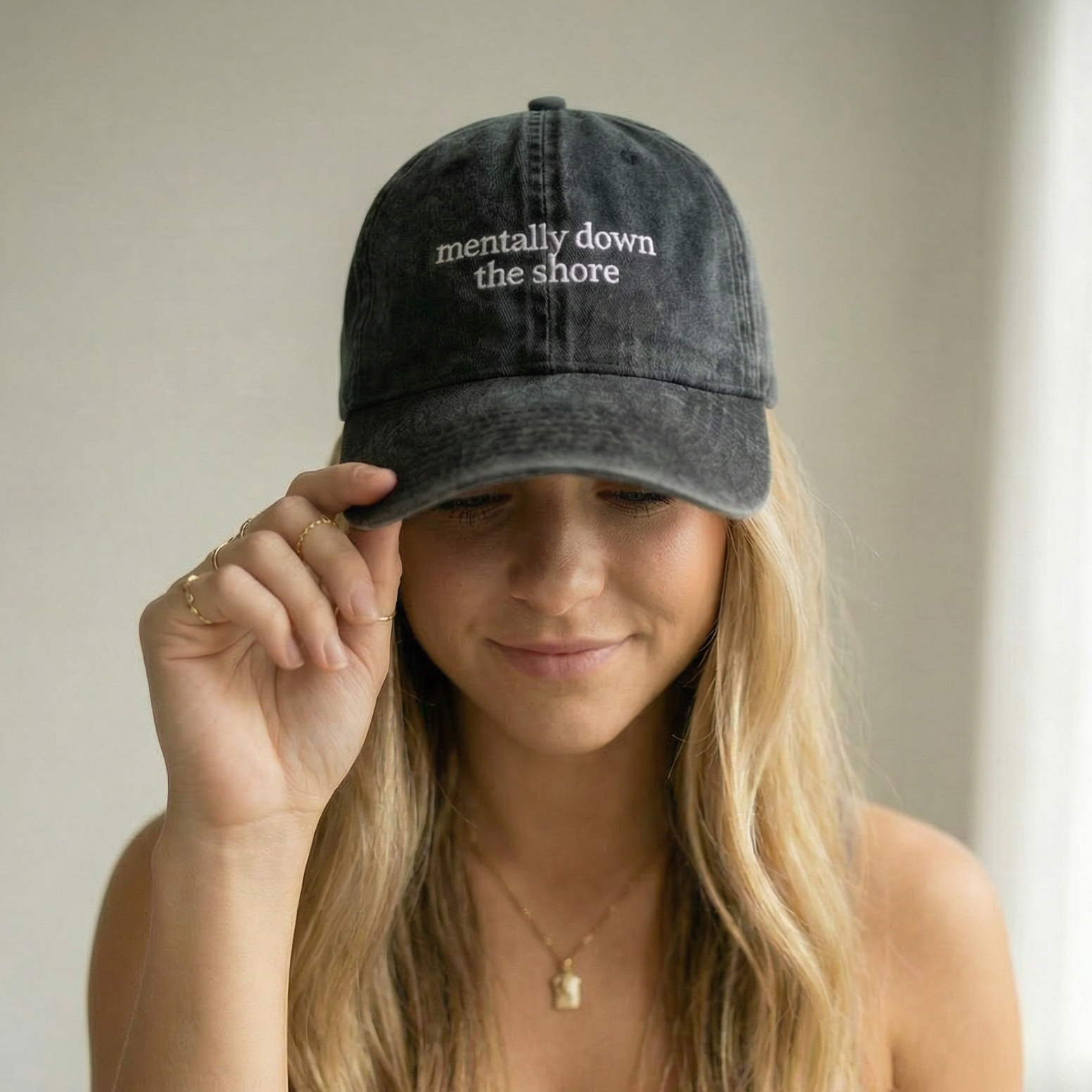Woman wearing a denim cap with 'mentally down the shore' text, holding it up to her face.