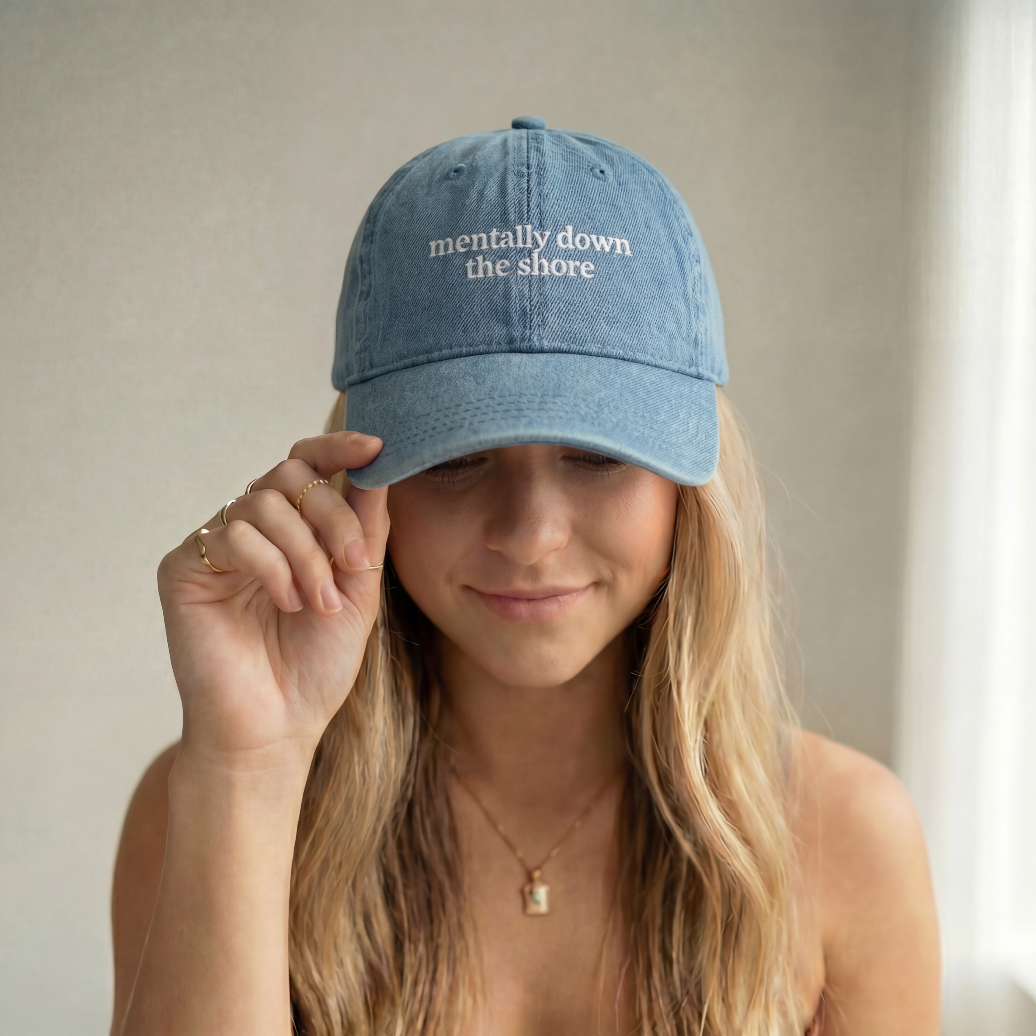 Woman wearing a blue cap that says "mentally down the shore" holding it on her head.